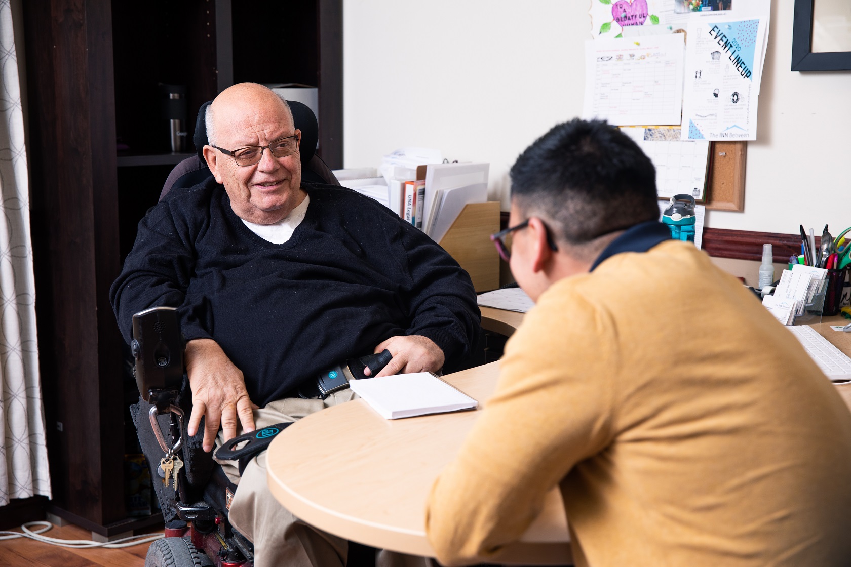 An elderly man in a wheelchair sitting at a desk with another man